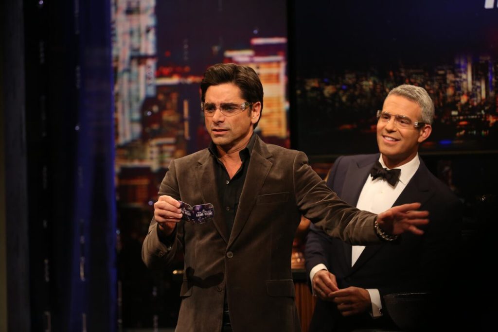 John Stamos gives a clue to his team as Andy Cohen looks on